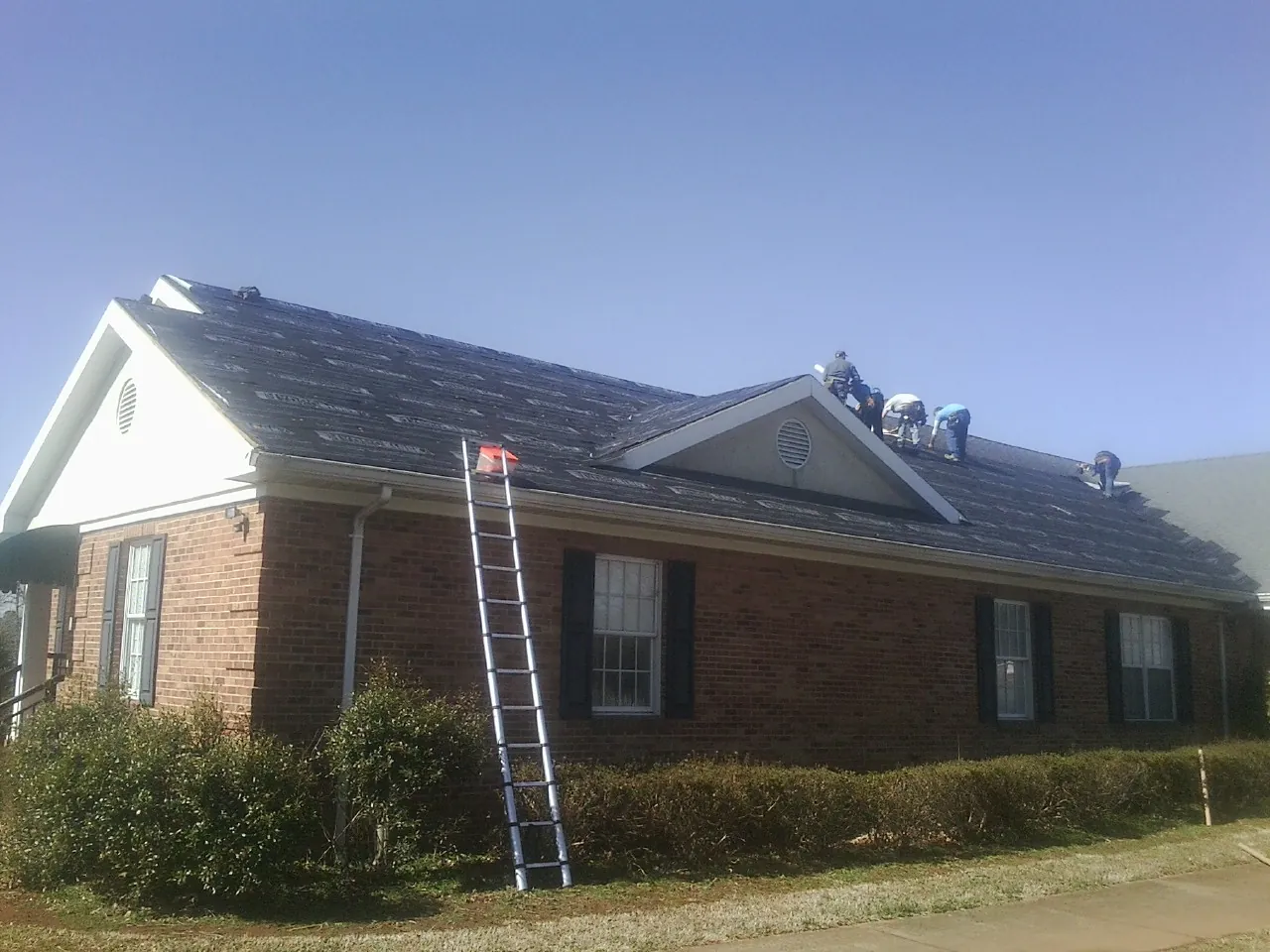Crew tearing shingles off a brick ranch roof with a ladder set — Metal Roof Repair work by our crew in Greenlawn