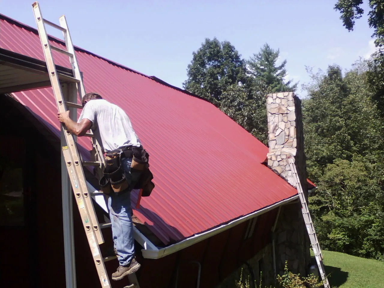 Roofer on a ladder beside a red metal roof and stone chimney — Soffit & Fascia Repair by Roofing Brothers in Greenlawn